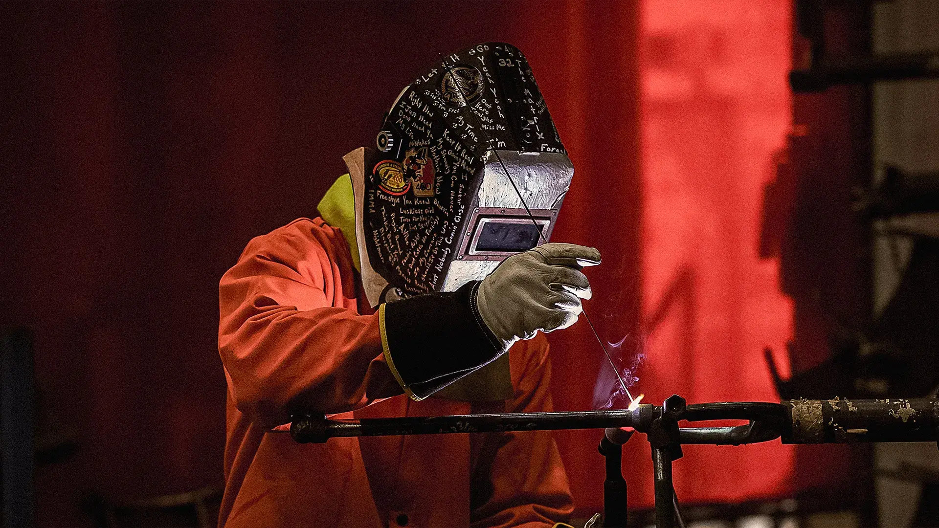 A man in a welding mask holds a metal rod, welding in a workshop environment.